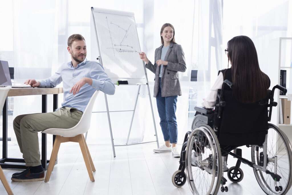 students in a classroom, one sits at a desk, one stand by a whiteboard, and one is a wheelchair user