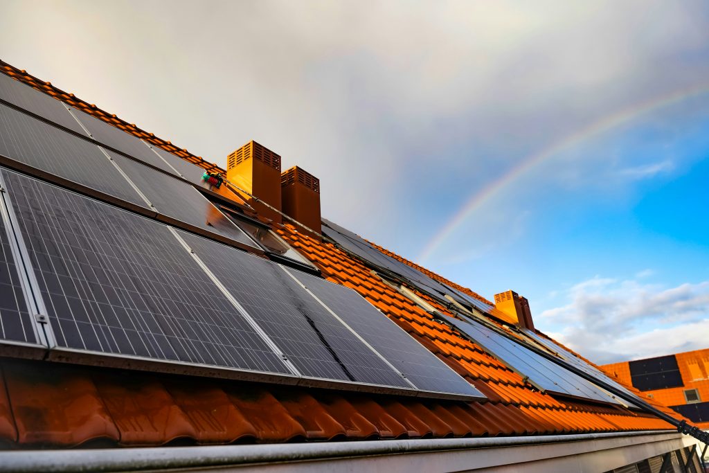 Solar panels installed on a sloped orange-tiled roof beneath a cloudy sky with a faint rainbow, symbolizing clean energy and a regenerative future.