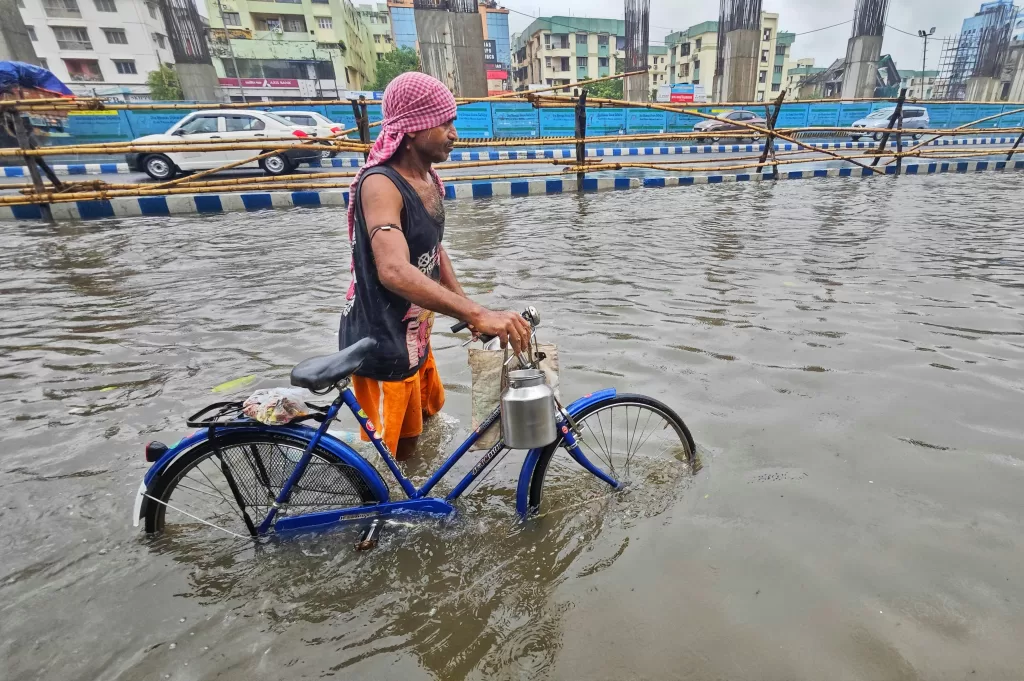 Man walking a bicycle through monsoon floodwaters in a city, highlighting the need for climate mitigation and adaptation in urban planning.
