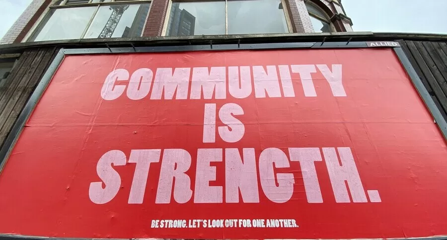 A large red billboard with bold white letters reading “COMMUNITY IS STRENGTH.” Smaller text below says, “Be strong. Let’s look out for one another.” The billboard is mounted on a wooden wall in front of a brick building.