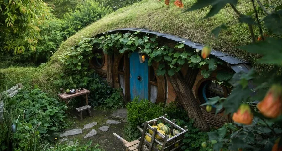 A cozy, hobbit-like house built into a hillside, featuring a grass-covered roof, round windows, and abundant edible plants surrounding the entrance.