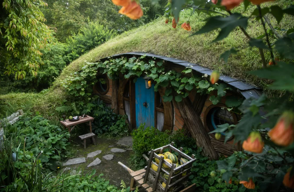 A cozy, hobbit-like house built into a hillside, featuring a grass-covered roof, round windows, and abundant edible plants surrounding the entrance.