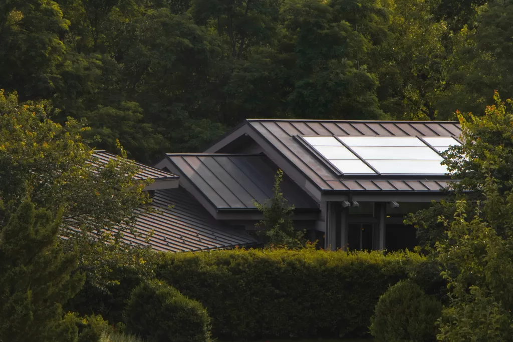 House with dark metal roofs and solar panels on top, set amid dense green trees and hedges.