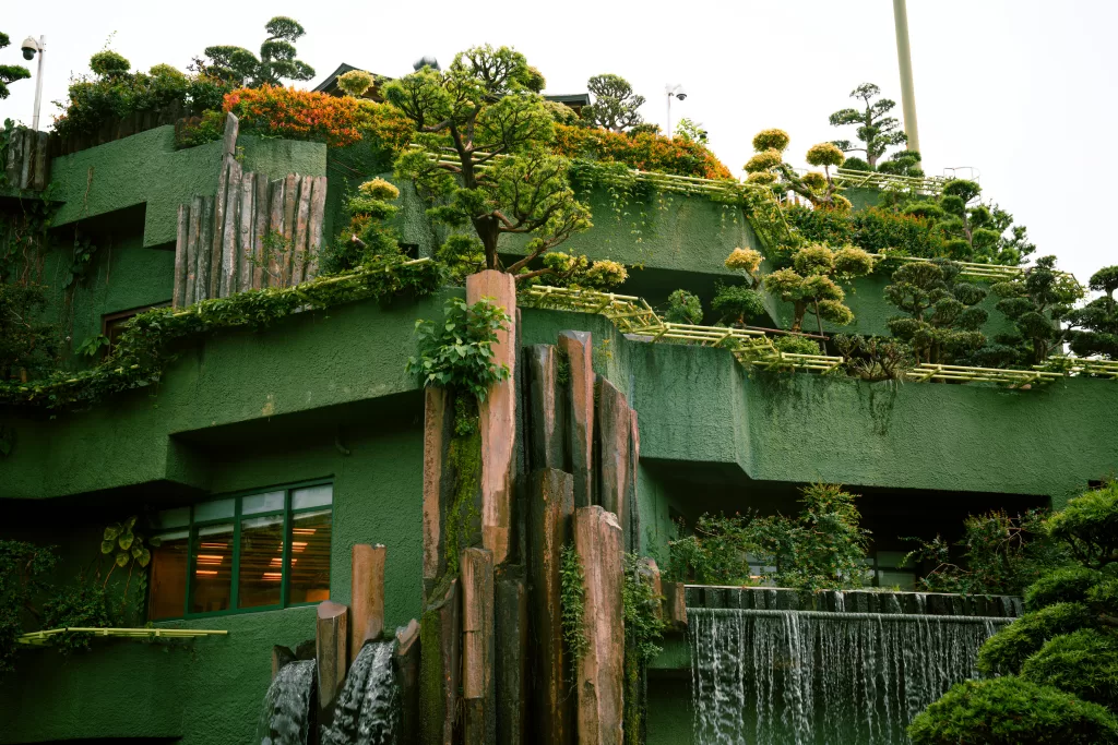 A terraced building with green walls, lush vegetation, bonsai-style trees, bamboo railings, and a waterfall feature flowing down the facade.