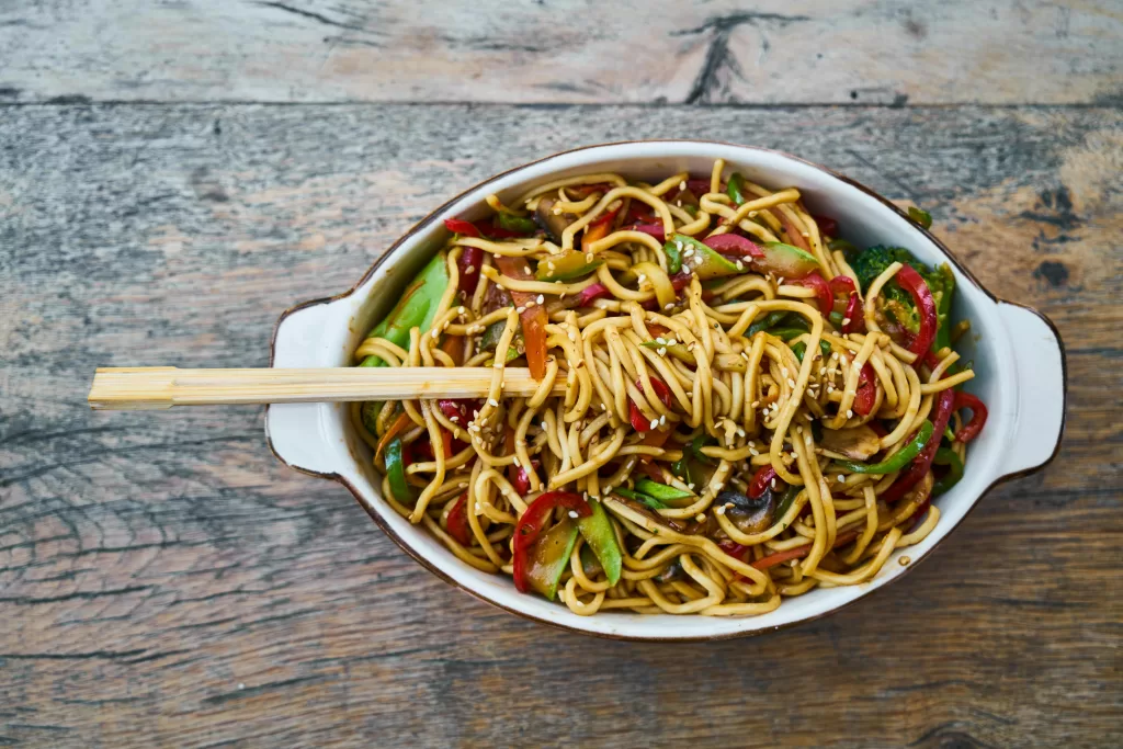 An oval dish sits on a table, full of noodles and vegetables. A pair of chopsticks are wrapped in the noodles.
